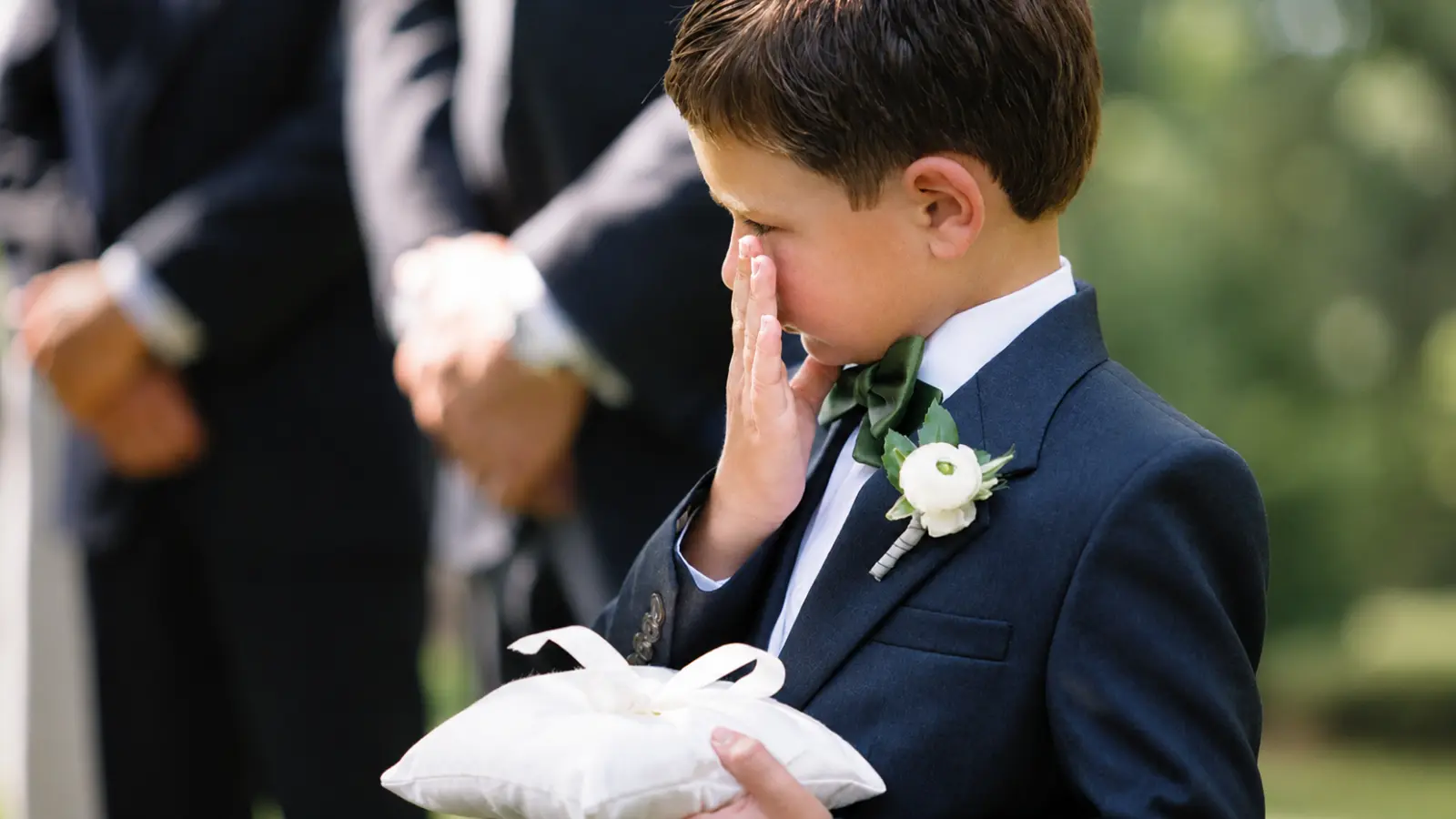Young ring bearer wearing a navy suit carries a white wedding ring pillow during the ceremony as part of his traditional ring bearer duties