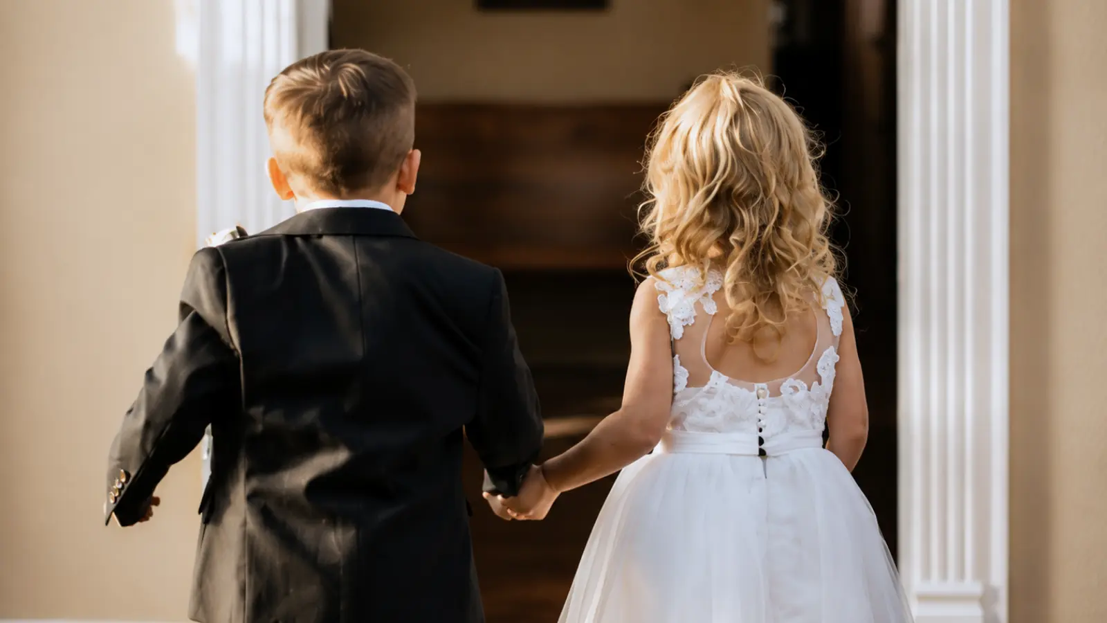 Ring bearer in a black suit and flower girl in a white dress walk hand in hand together during the wedding ceremony processional