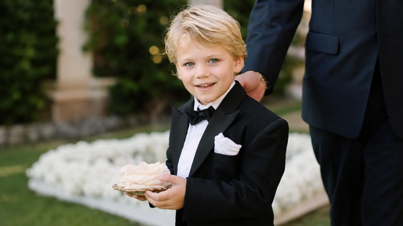 Young ring bearer dressed in a classic black suit with white shirt, black bow tie and white pocket square at an outdoor wedding