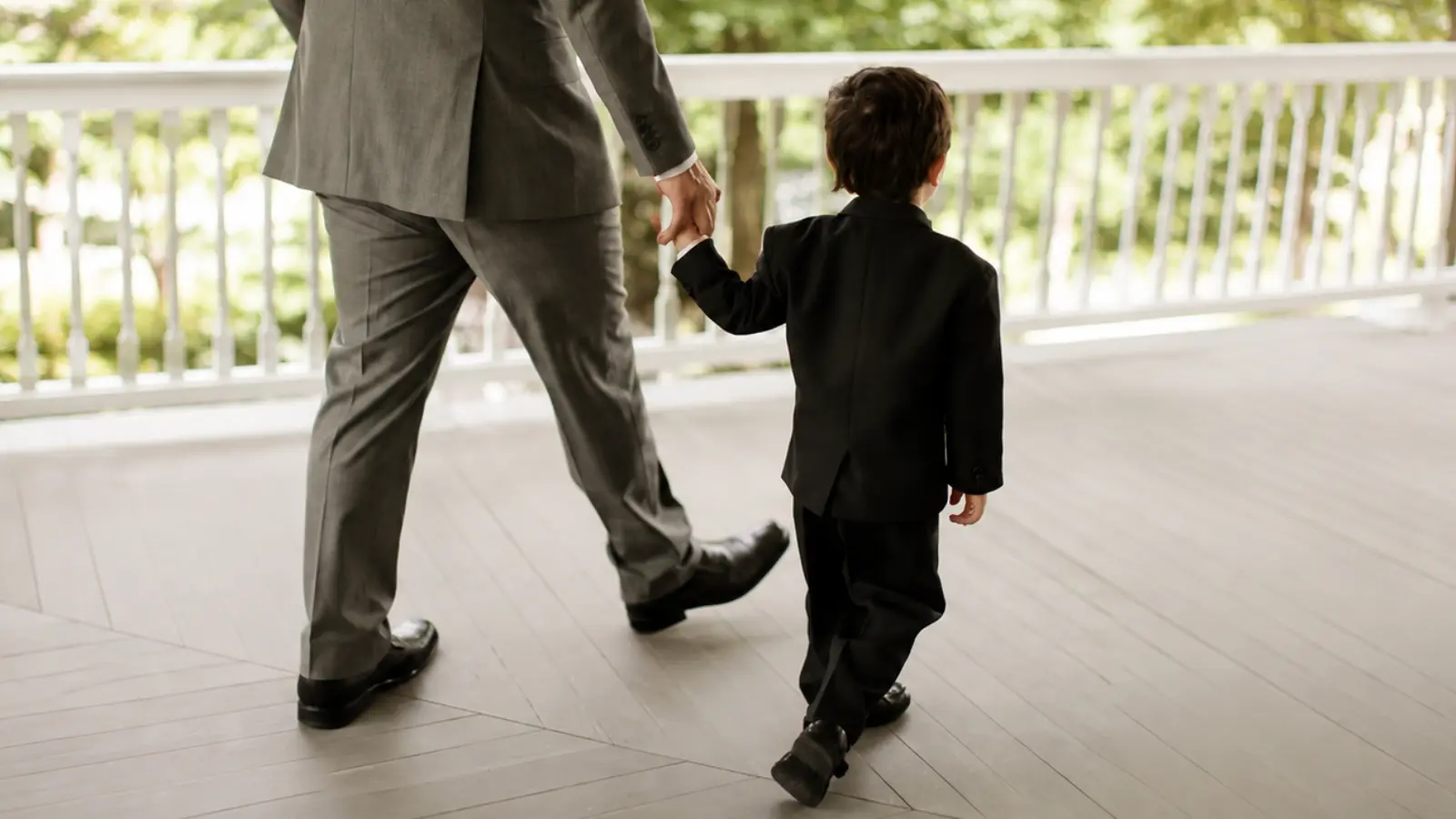 Small ring bearer wearing a black suit walks with an adult holding his hand for guidance and reassurance during the wedding ceremony processional
