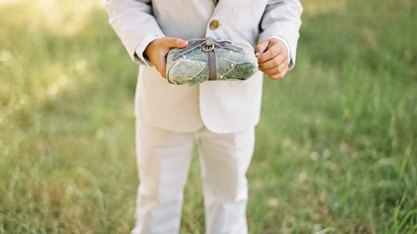 Young ring bearer wearing a light suit and carrying a decorative green wedding ring pillow as a practical ceremony gift
