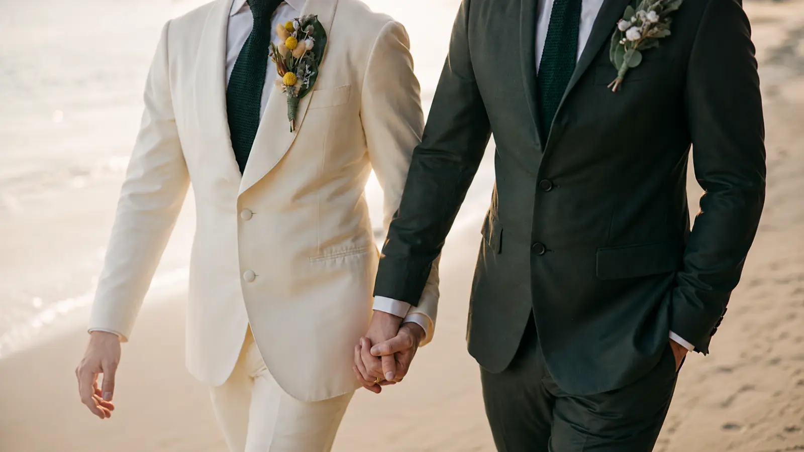 Same-sex wedding couple holding hands in cream and dark green suits with boutonnieres during an outdoor beach wedding celebration Same-Sex Wedding Planning: Traditions and Inclusive Logistics
