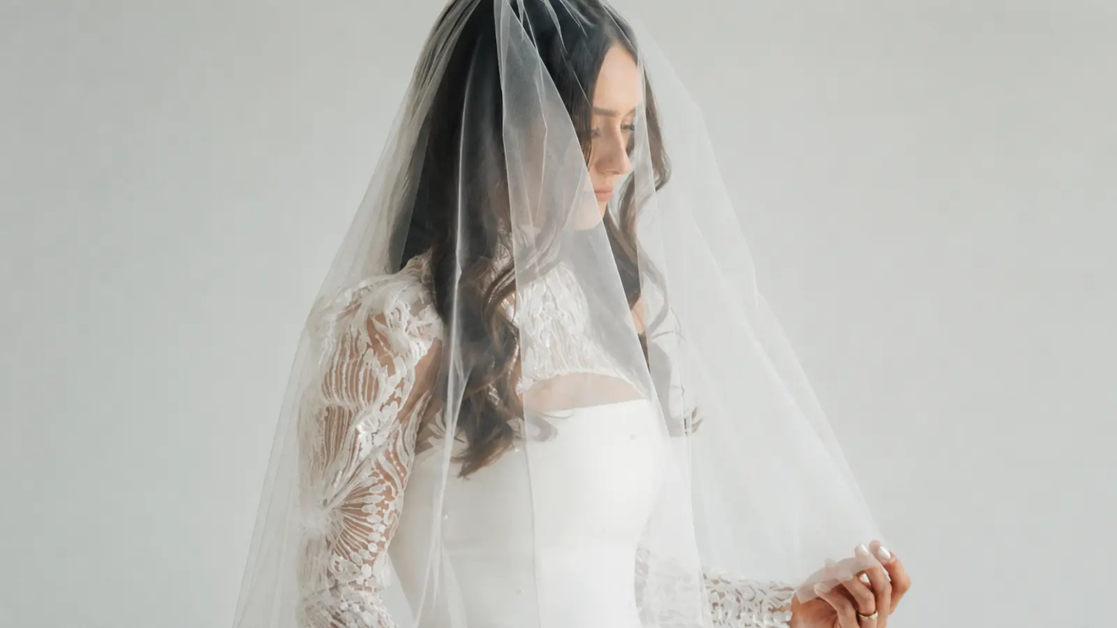 Bride wearing a long sheer blusher wedding veil over her face with a white lace sleeve wedding dress and softly styled dark hair