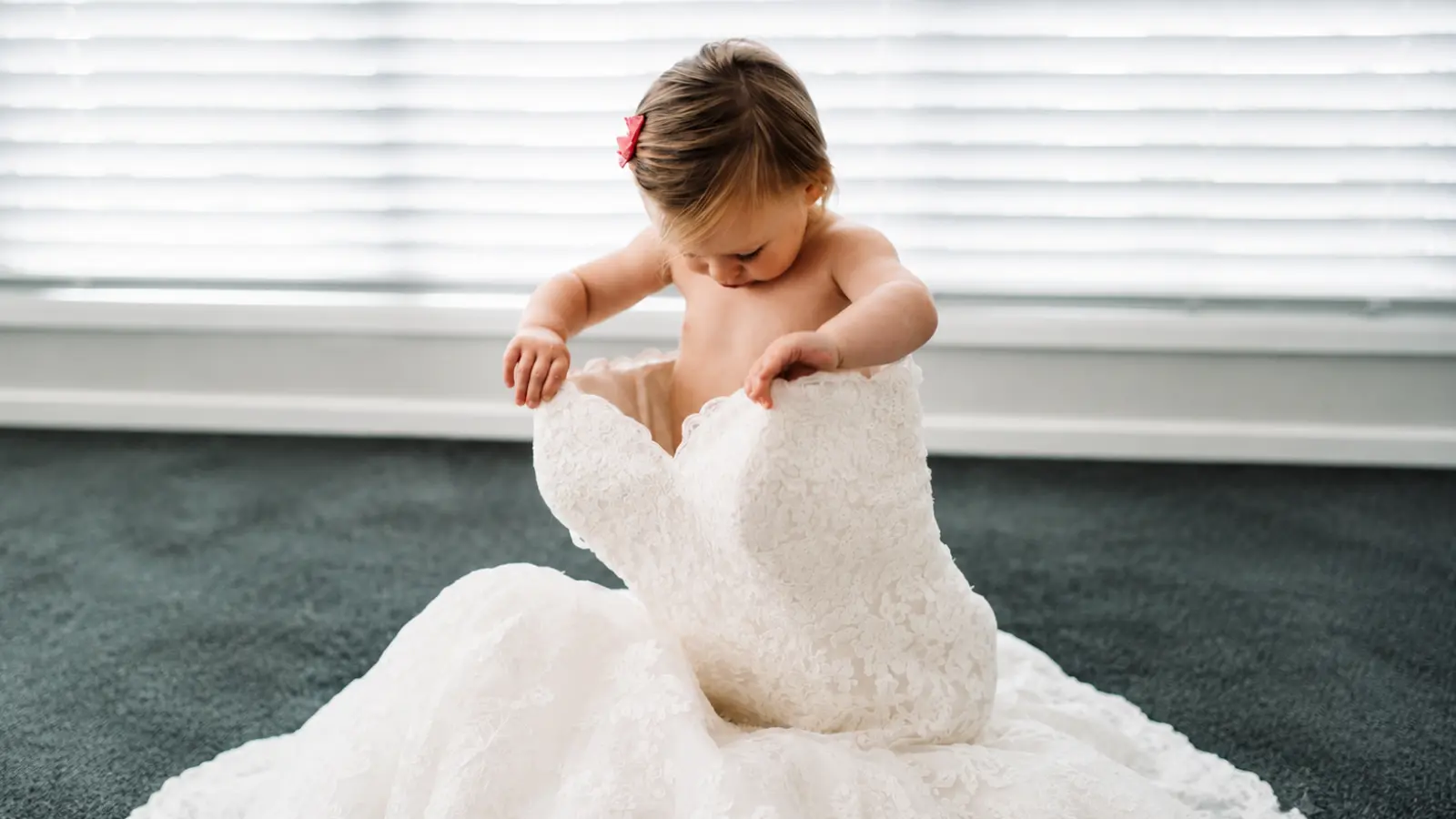 Young toddler sits on the floor while exploring a white lace flower girl dress before a wedding celebration Flower Girl Age