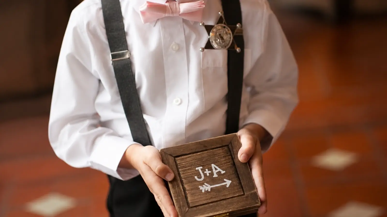 Ring bearer holding a personalized wooden box with couple initials while wearing suspenders and a pink bow tie during the wedding ceremony Ring Bearer Ideas