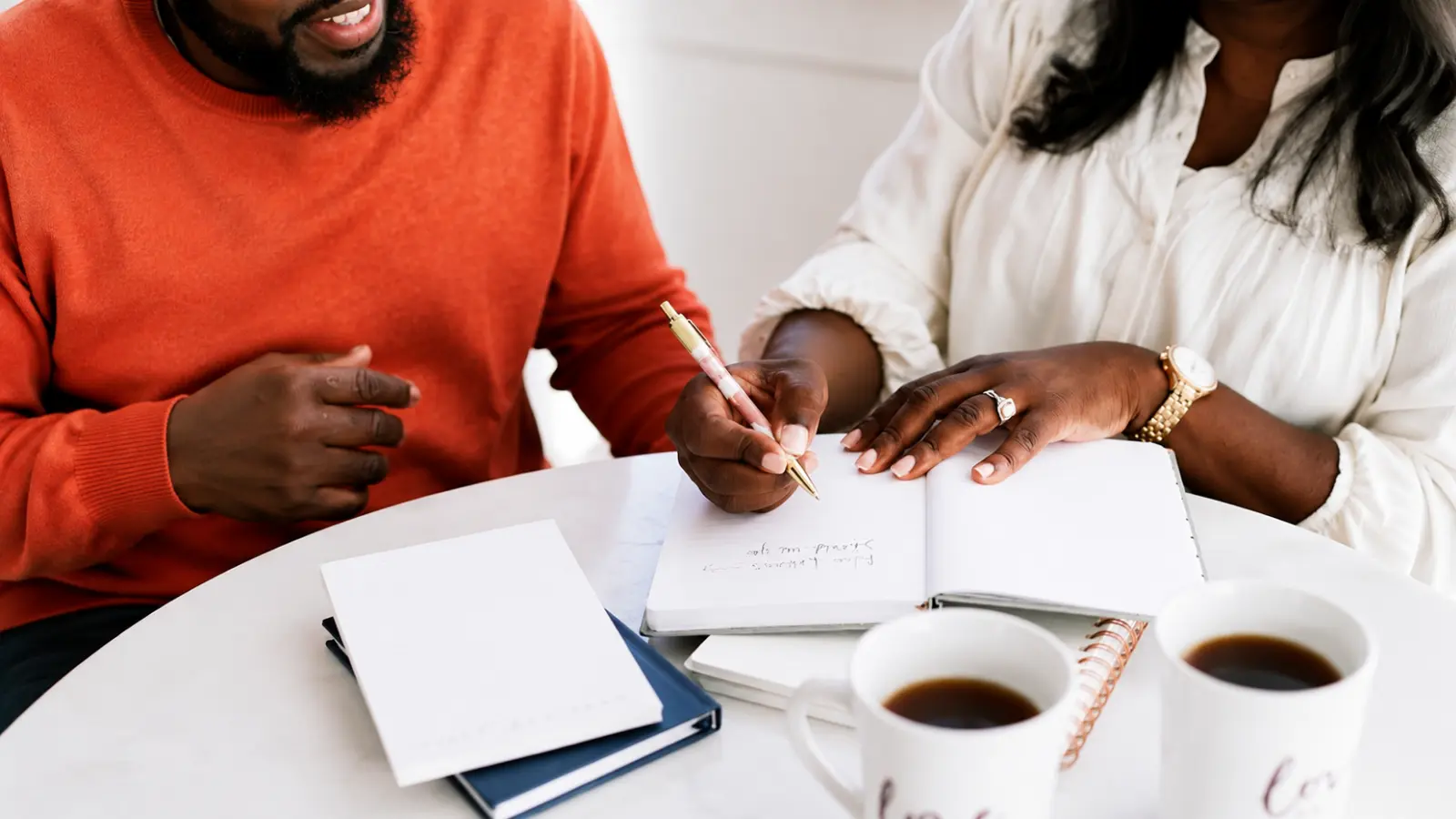Engaged couple writing wedding planning notes together at a table with notebooks and coffee while discussing ideas and decisions for their wedding