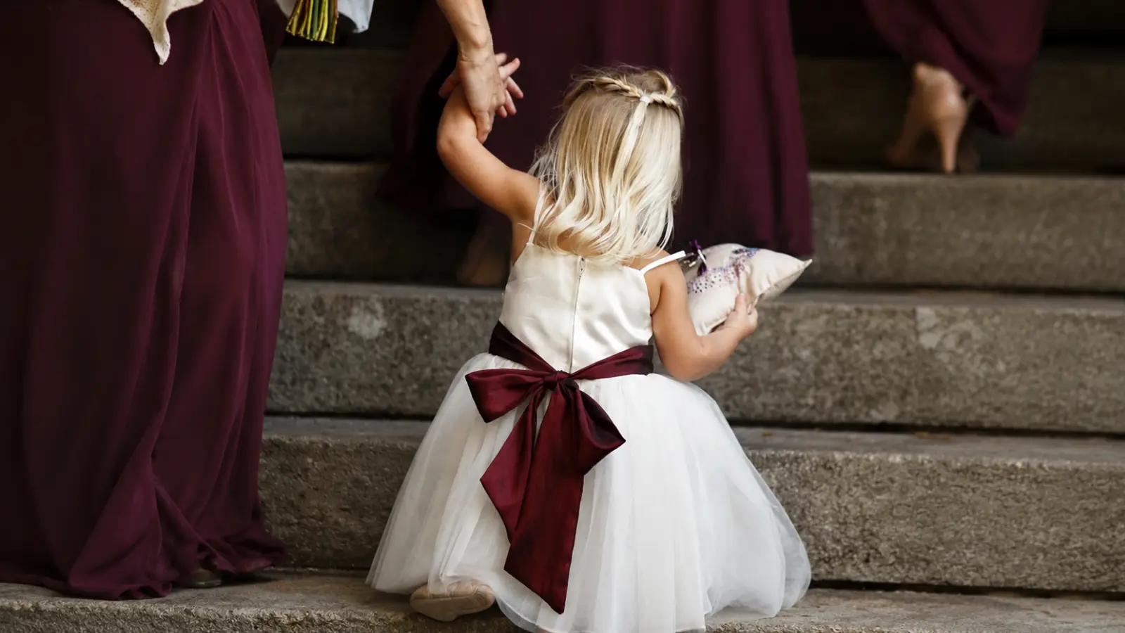 Young flower girl wears a white wedding dress with a large burgundy sash and carries a small ring pillow while walking upstairs