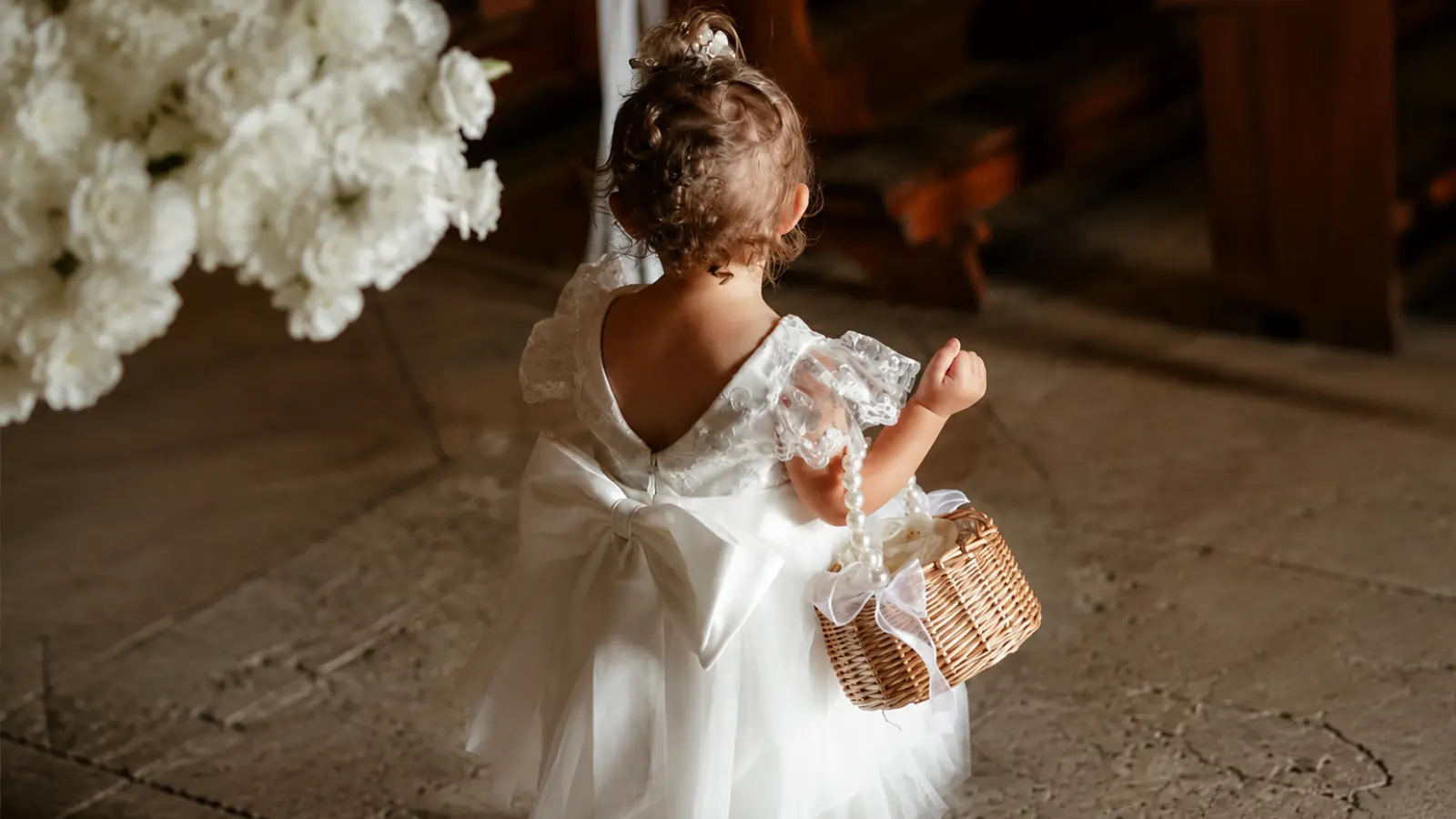 Young flower girl wears a white dress with lace sleeves and large back bow while carrying a woven flower basket at a wedding