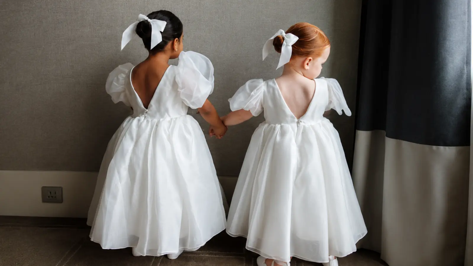 Two flower girls hold hands wearing matching white dresses with puff sleeves, full skirts and coordinating white bows in their hair Flower Girl Dresses