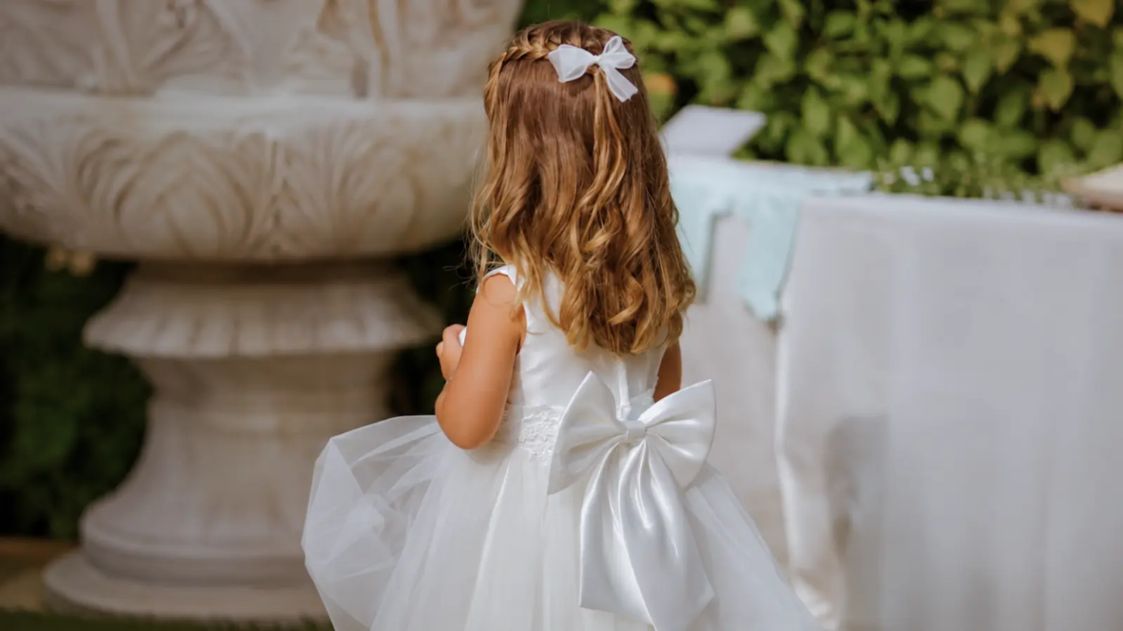 Young flower girl wears a white tulle dress with an oversized satin bow while standing at an outdoor wedding ceremony