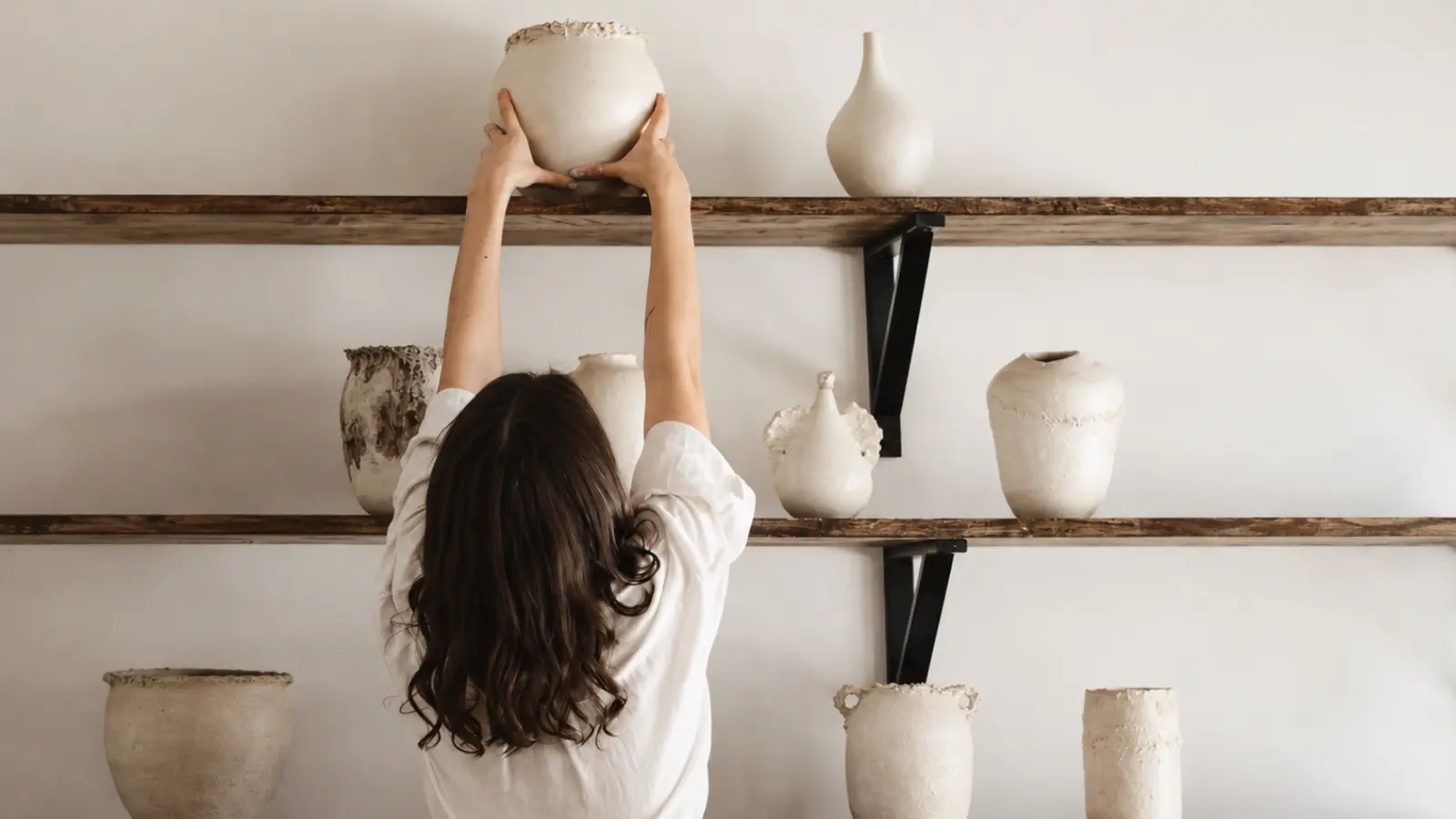 Woman arranging handmade ceramic vessels on wooden shelves as decorative pottery gift ideas for an 8th wedding anniversary
