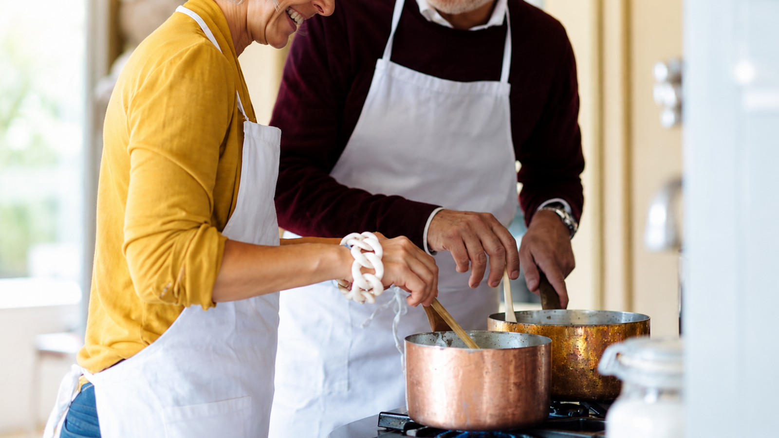 Older couple cooking together in a bright home kitchen, stirring food in copper-colored saucepans while wearing white aprons, illustrating a shared culinary experience inspired by 22nd wedding anniversary copper gifts