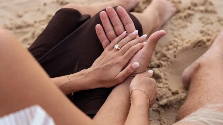 Close-up of a couple sitting together on a sandy beach with their hands gently resting against each other, a sparkling gemstone ring and delicate bracelets visible, representing the lasting love and commitment celebrated on a 25th wedding anniversary